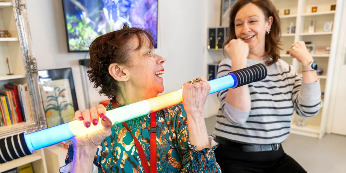 Twee vrouwen lachen terwijl ze samen met een felgekleurde oefenstang bezig zijn. Ze zijn in een lichte kamer met een televisie op de achtergrond.