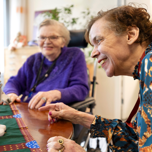 Twee lachende vrouwen zitten aan een tafel en kijken naar een kleine robot. Een van hen wijst er enthousiast naar.